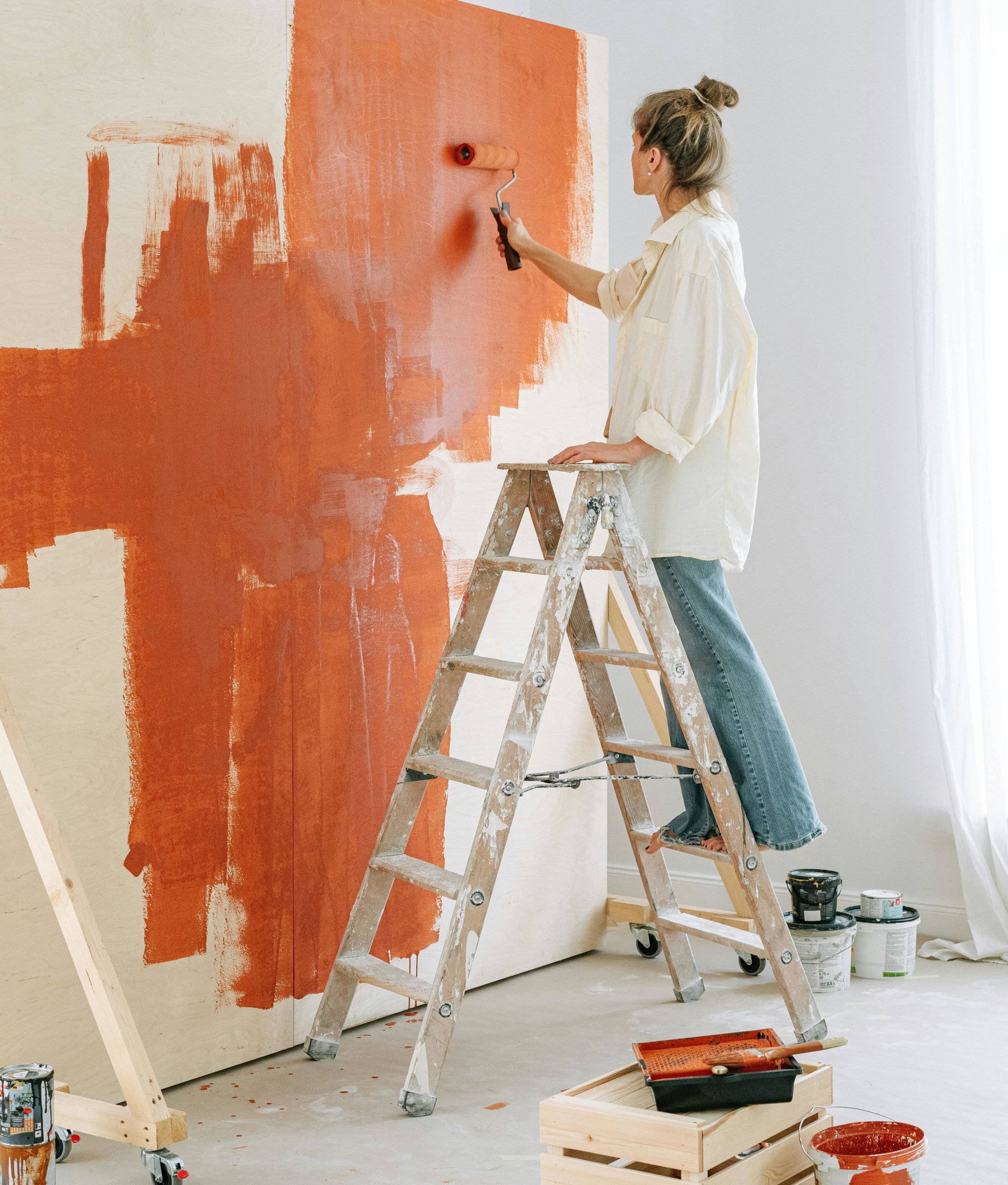 Woman on a stepladder painting a wall a burnt orange colour with a paint roller