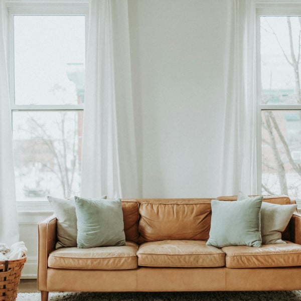 Windows with white curtains behind a brown leather sofa with cushions